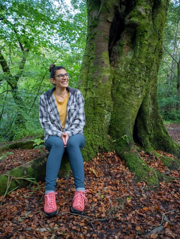 Francesca is sitting on a tree root, wearing colourful hiking clothing, and smiling at her environment. The outdoor therapist is in a woodland area, where there are a lot of dry leaves on the ground.